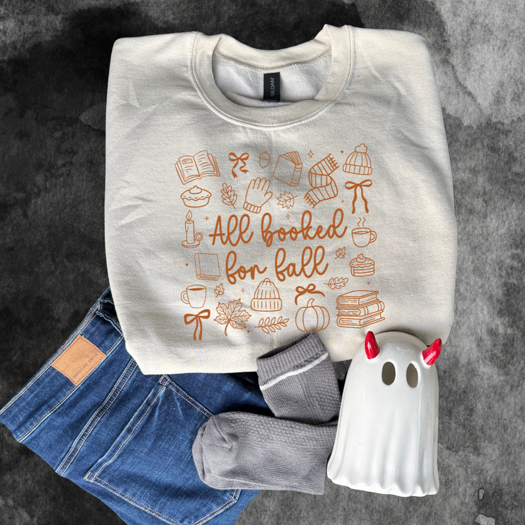 Flat lay of a sweatshirt with 'All booked for fall' text and graphics, jeans, socks, and ghost decoration on a textured surface by Fiction & Fibre.