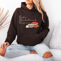 A woman wearing a dark brown hoodie with a graphic that reads 'Books Blankets and Bonfires' above an illustration of stacked books, a cup, and a fire, paired with light-wash ripped jeans, sitting on a white couch.