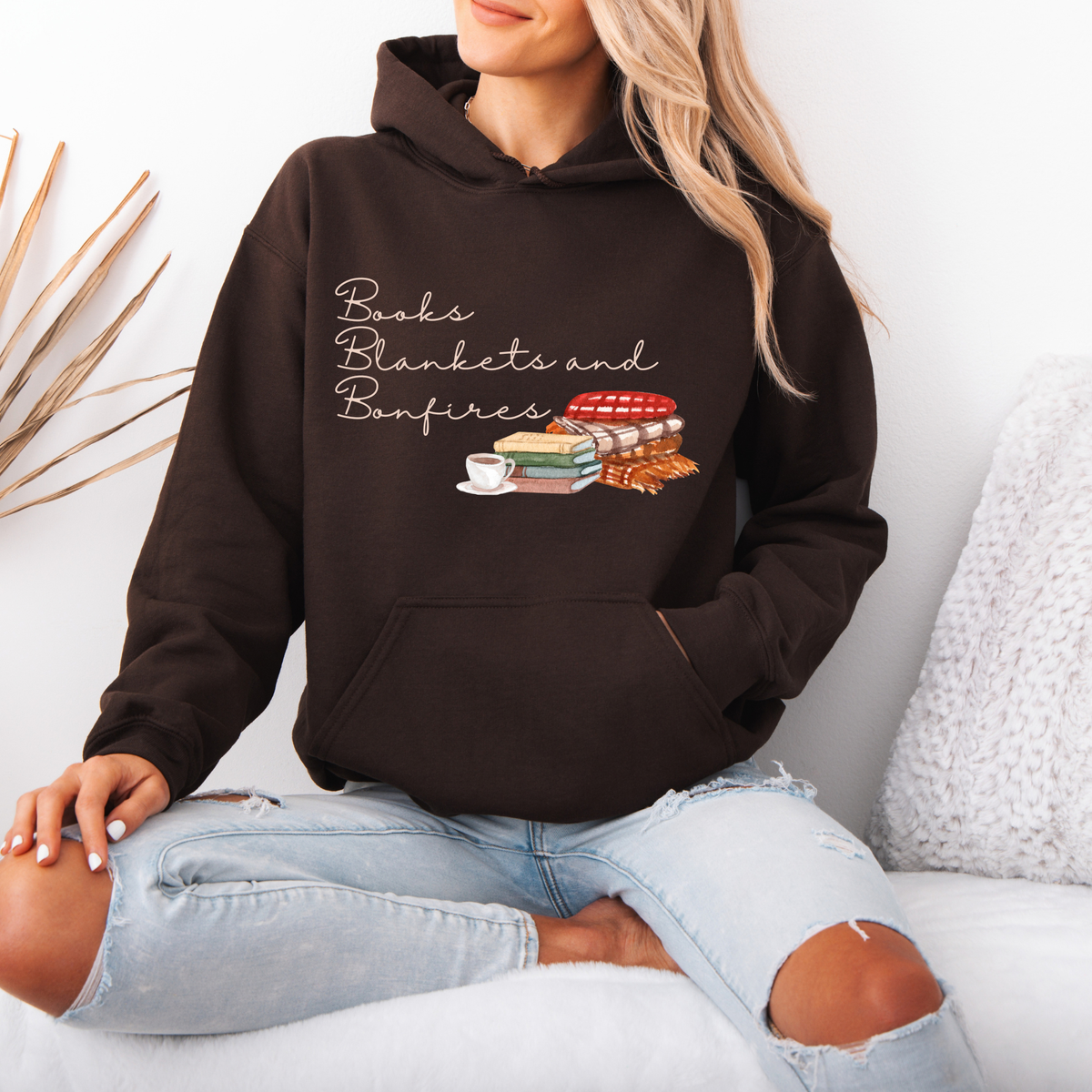 A woman wearing a dark brown hoodie with a graphic that reads 'Books Blankets and Bonfires' above an illustration of stacked books, a cup, and a fire, paired with light-wash ripped jeans, sitting on a white couch.