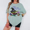 A woman wearing an oversized sage green graphic t-shirt with a 'Books, Brews and Witchy Views' design featuring a witch hat, books, crystals, and a drink, paired with black bike shorts.