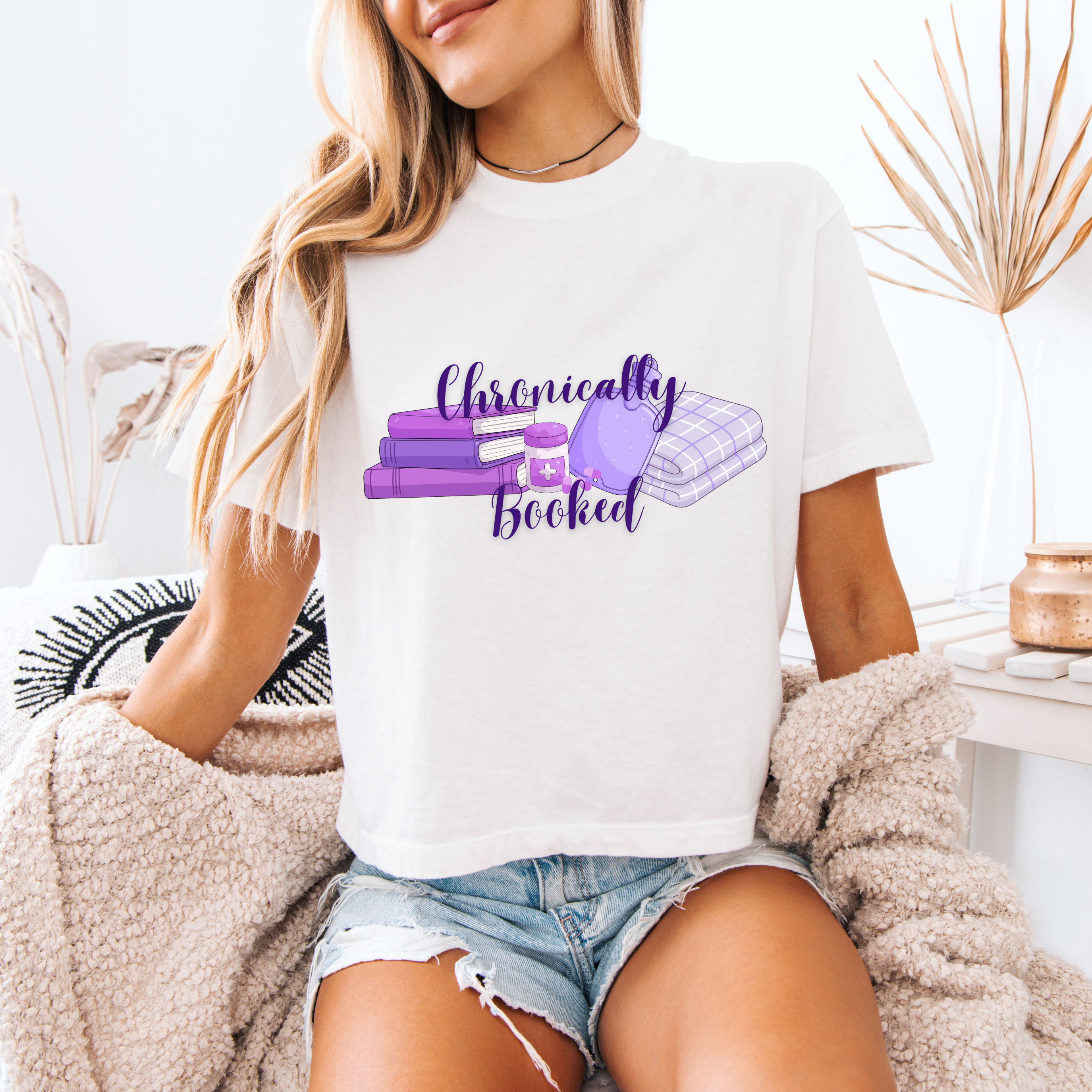 A woman wearing a white boxy crop that reads "chronically booked" with a stack of books, medicine, a hot water bottle and a blanket.