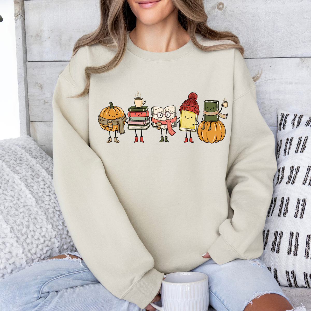 A woman wearing a cream-colored crewneck sweatshirt with a colorful graphic of autumn-themed characters (a pumpkin, books, a coffee cup, a book, a hot air balloon, and a pumpkin) sitting in a cozy room setting.
