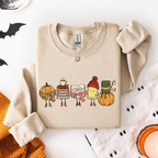 A folded beige sweatshirt with a colorful graphic of five cute, cartoonish characters dressed in autumn and winter clothing, displayed on a white flat-lay background with Halloween-themed props.