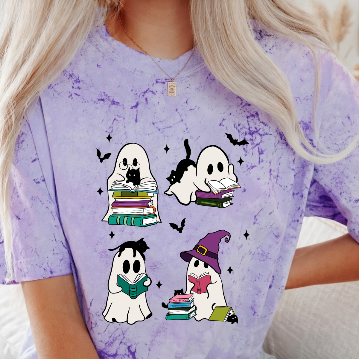 A person wearing a light purple tie-dye t-shirt with a cartoon graphic of four ghosts reading books in a Halloween theme. The person has blonde hair and is wearing a necklace.