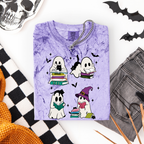 A folded lavender-purple t-shirt with a Halloween graphic print of four cute ghosts reading books, styled in a flat-lay with a skull, black-and-white checkered fabric, gray jeans, and orange pencils on a white background.