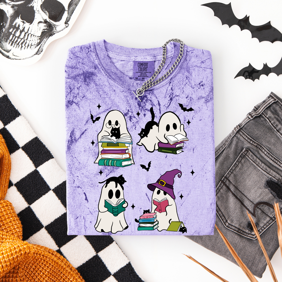 A folded lavender-purple t-shirt with a Halloween graphic print of four cute ghosts reading books, styled in a flat-lay with a skull, black-and-white checkered fabric, gray jeans, and orange pencils on a white background.