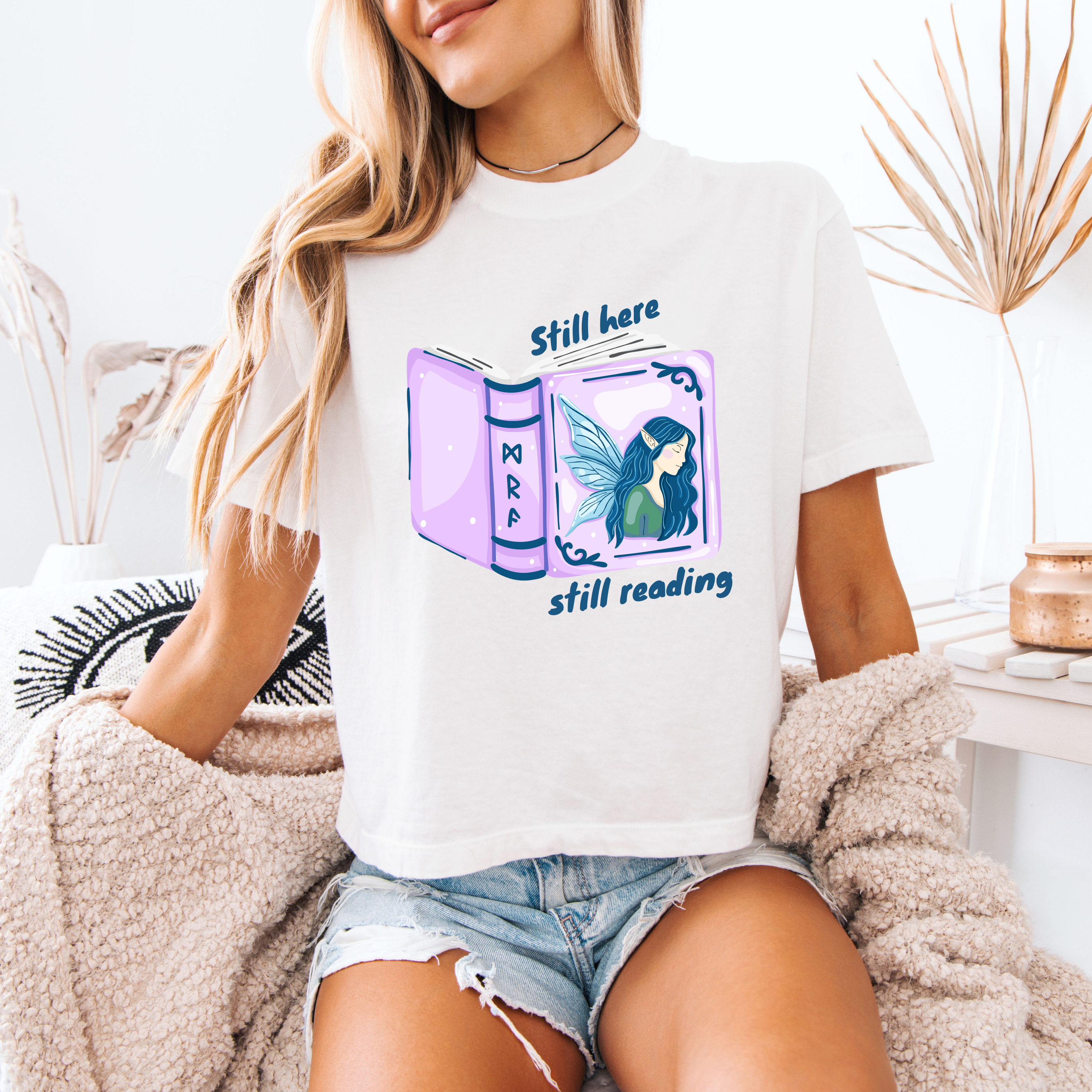 A woman wearing a white boxy crop that reads "still here still reading" with an open book on the front that has a fairy on it