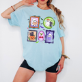 A woman wearing a light blue graphic T-shirt with Halloween-themed framed illustrations and black shorts, posed against a plain white background.