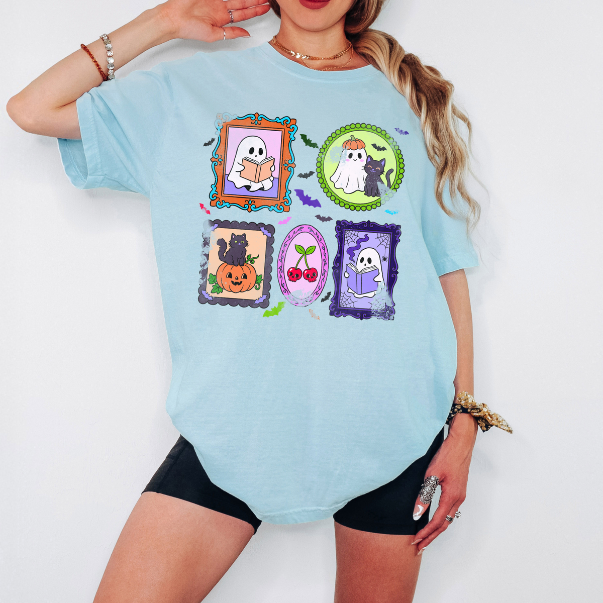 A woman wearing a light blue graphic T-shirt with Halloween-themed framed illustrations and black shorts, posed against a plain white background.