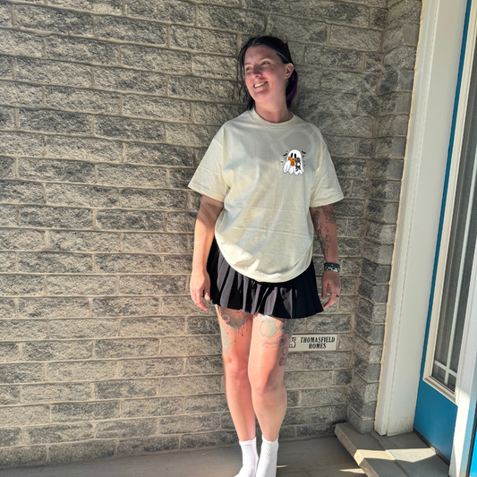 Person wearing a light-colored t-shirt with a Fiction & Fibre design, ghost and cats, black shorts, and white socks standing against a brick wall.