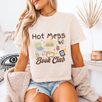 A woman wearing a cream-coloured graphic t-shirt with 'Hot Mess Book Club' printed in brown text surrounded by colourful book-themed doodles. She is sitting in a cozy, modern room with a beige sherpa cardigan and denim shorts.