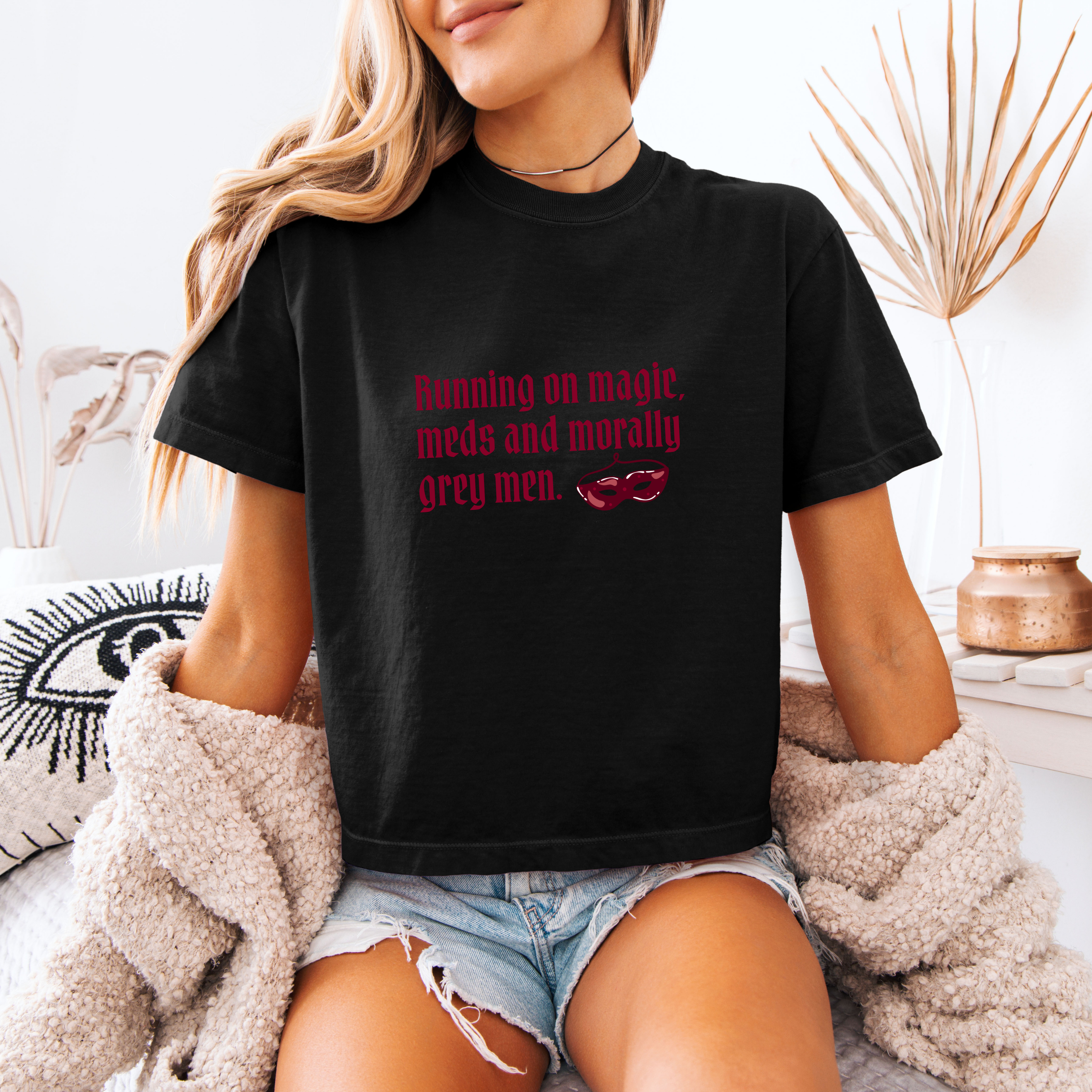 A woman wearing a black boxy crop tee that has a crown and reads "Running on magic, meds and morally grey men"