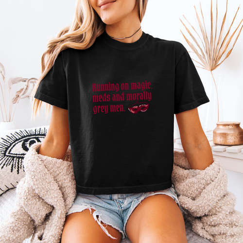 A woman wearing a black boxy crop tee that has a crown and reads "Running on magic, meds and morally grey men"