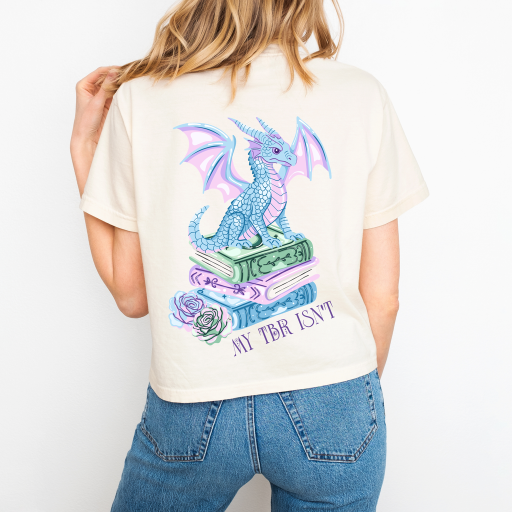 A woman wearing an Ivory boxy crop that shows the back and reads "My TBR isnt" and has a dragon sitting on a stack of books