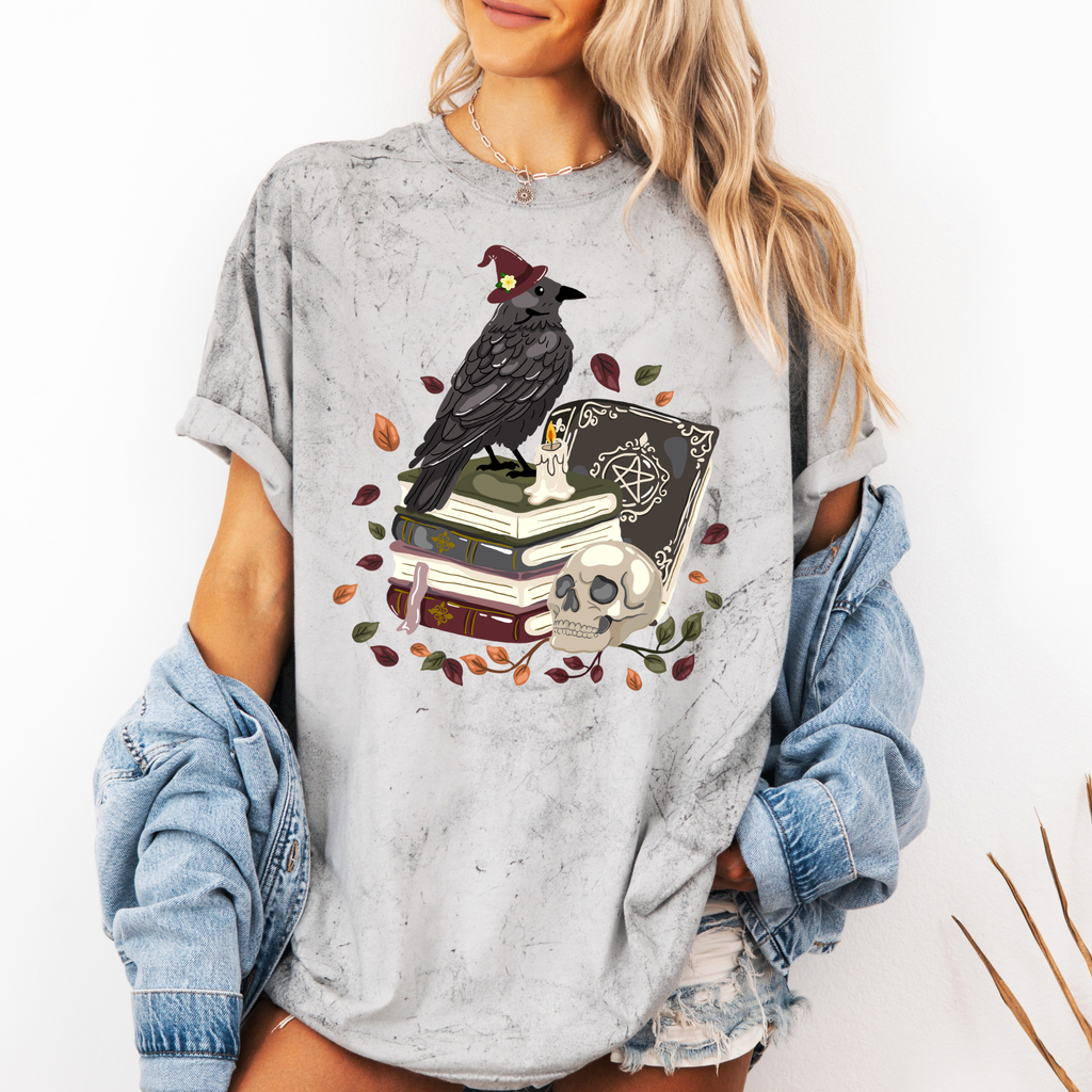 A woman wearing a light gray distressed graphic t-shirt featuring a black raven on stacked books with a skull and autumn leaves. She has a light blue denim jacket off her shoulders and is posing against a white wall.