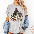 A woman wearing a light gray distressed graphic t-shirt featuring a black raven on stacked books with a skull and autumn leaves. She has a light blue denim jacket off her shoulders and is posing against a white wall.
