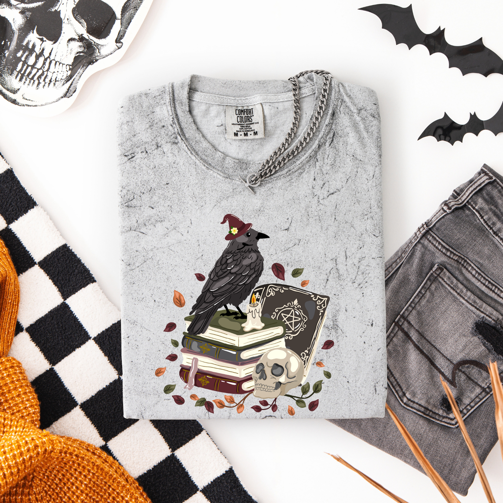 A folded light gray graphic t-shirt with a gothic raven design, styled in a flat lay with gray jeans, a black and white checkered blanket, a skull, and bat cutouts on a white background.