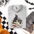 A folded light gray graphic t-shirt with a gothic raven design, styled in a flat lay with gray jeans, a black and white checkered blanket, a skull, and bat cutouts on a white background.