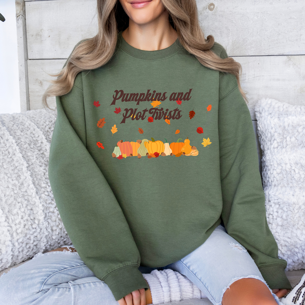 A woman wearing an olive green crewneck sweatshirt with a 'Pumpkins and Plot Twists' autumn graphic, sitting on a cozy white couch.
