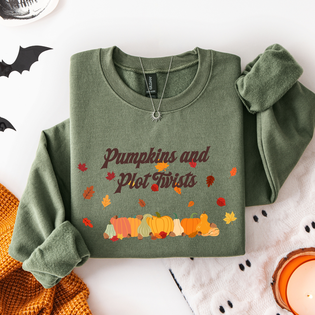 An olive green crewneck sweatshirt with a 'Pumpkins and Plot Twists' graphic featuring autumn leaves and pumpkins, styled in a cozy flat-lay setting with a candle, knit blanket, and black bat cutouts.