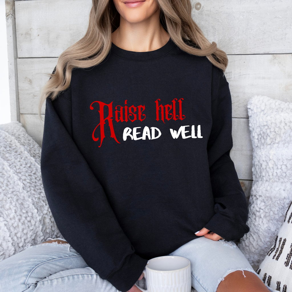 A woman wearing a black crewneck sweatshirt with the text 'Raise hell READ WELL' printed on the front in red and white, paired with light blue ripped jeans, sitting on a couch with a white mug.