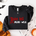 A folded black sweatshirt with the text 'Raise hell READ WELL' printed on the front in red and white, styled in a flat lay with Halloween-themed props including bat cutouts, a knit orange sweater, a white fabric with black dots, and a lit candle.