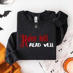 A folded black sweatshirt with the text 'Raise hell READ WELL' printed on the front in red and white, styled in a flat lay with Halloween-themed props including bat cutouts, a knit orange sweater, a white fabric with black dots, and a lit candle.