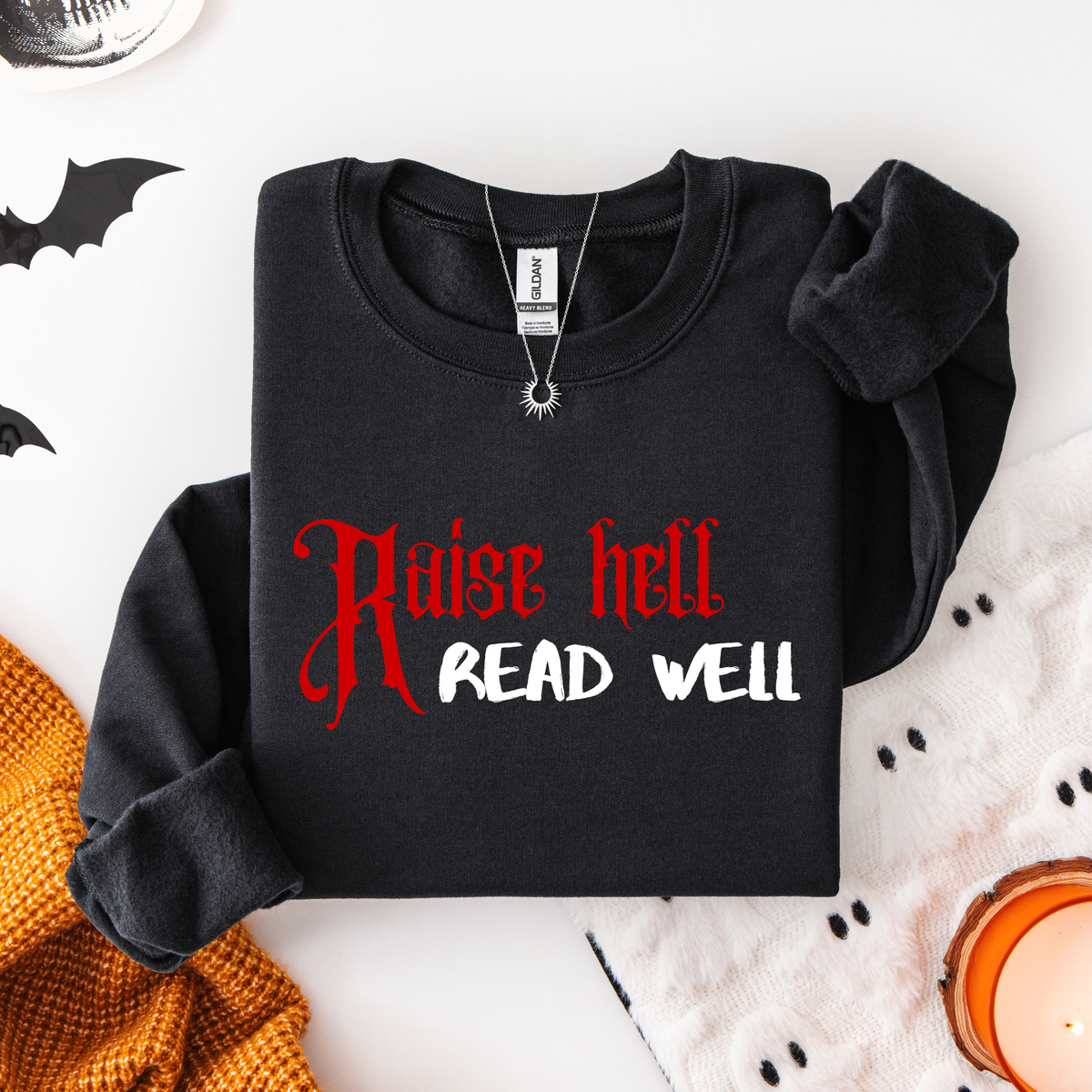 A folded black sweatshirt with the text 'Raise hell READ WELL' printed on the front in red and white, styled in a flat lay with Halloween-themed props including bat cutouts, a knit orange sweater, a white fabric with black dots, and a lit candle.