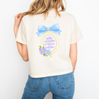 A woman wearing an Ivory boxy crop that shows the back reads "Darling keep blooming you are doing beautifully" in an oval frame with a bow