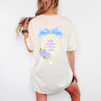 A woman wearing an ivory tee that is showing the back and has a frame with a bow that reads "darling keep blooming, you are doing beautifully" 
