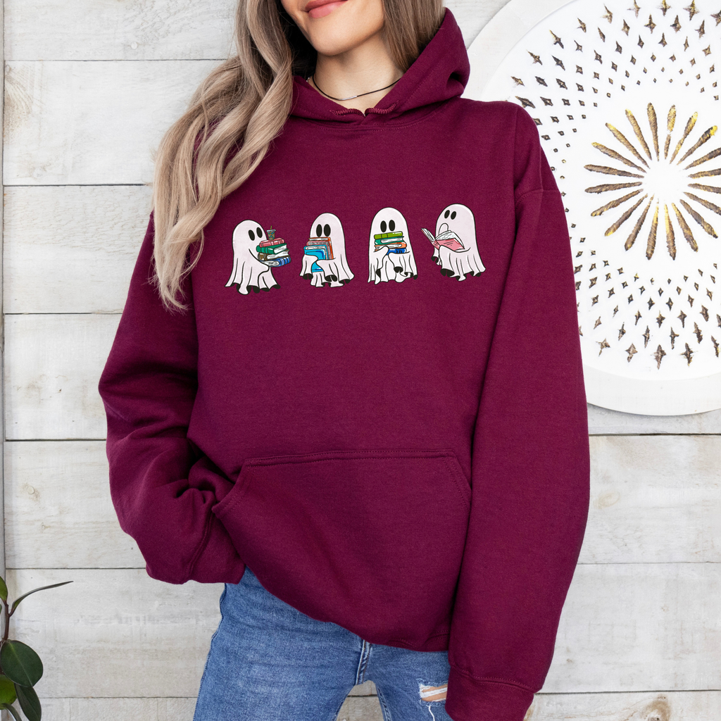 A woman wearing a maroon hoodie with a graphic of five cartoon ghosts holding colorful books, paired with blue jeans, standing against a white wood-paneled wall.
