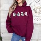 A woman wearing a maroon hoodie with a graphic of five cartoon ghosts holding colorful books, paired with blue jeans, standing against a white wood-paneled wall.