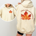 A cream-colored hoodie shown on a model, featuring a large orange and red autumn leaf graphic on the back with the text 'Leaves, Lattes and Local Books' in brown script. An inset circle shows the front view with a smaller matching leaf graphic on the chest.