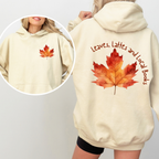 A cream-colored hoodie shown on a model, featuring a large orange and red autumn leaf graphic on the back with the text 'Leaves, Lattes and Local Books' in brown script. An inset circle shows the front view with a smaller matching leaf graphic on the chest.