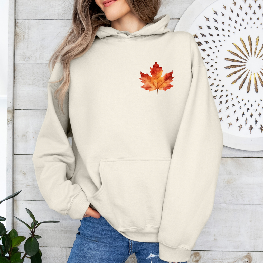 A woman wearing a cream-colored hoodie with a red and orange maple leaf graphic on the chest, paired with blue jeans, standing against a white wood-paneled wall.