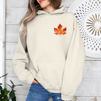 A woman wearing a cream-colored hoodie with a red and orange maple leaf graphic on the chest, paired with blue jeans, standing against a white wood-paneled wall.