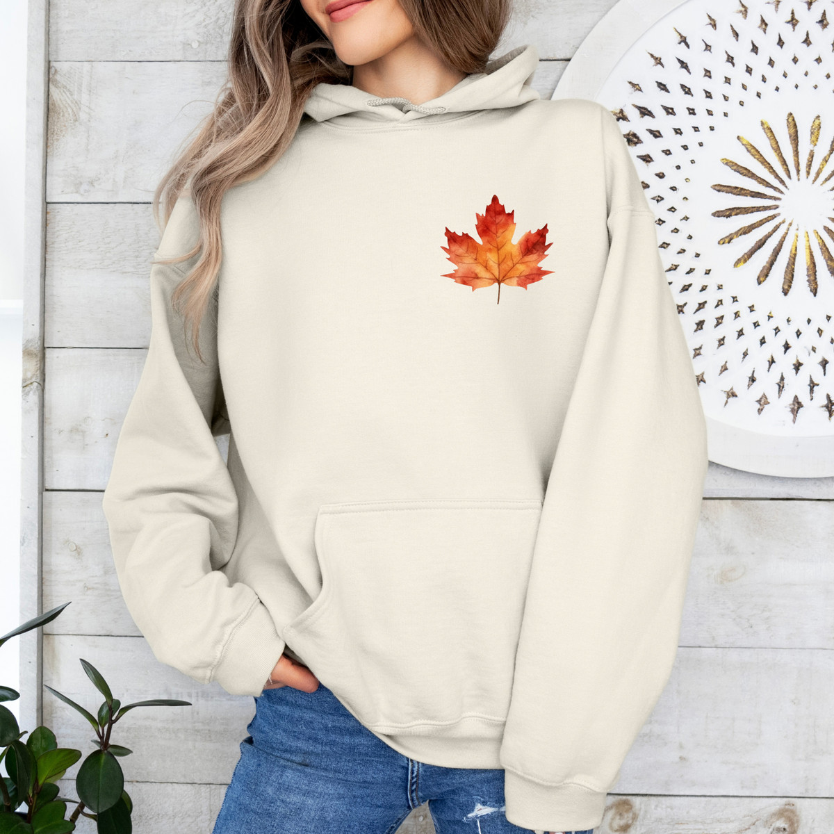 A woman wearing a cream-colored hoodie with a red and orange maple leaf graphic on the chest, paired with blue jeans, standing against a white wood-paneled wall.