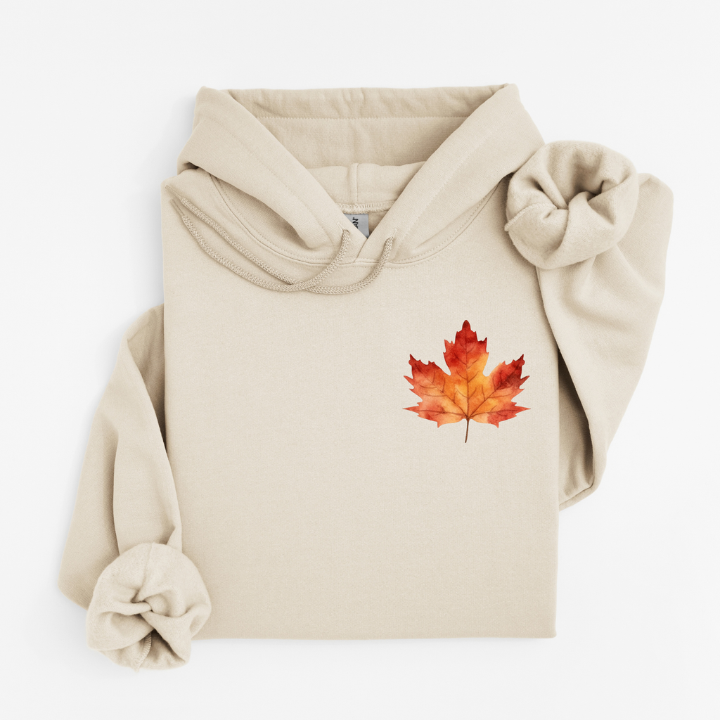 A beige pullover hoodie with a watercolor-style red and orange maple leaf graphic on the left chest, styled flat on a white background.