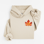 A beige pullover hoodie with a watercolor-style red and orange maple leaf graphic on the left chest, styled flat on a white background.